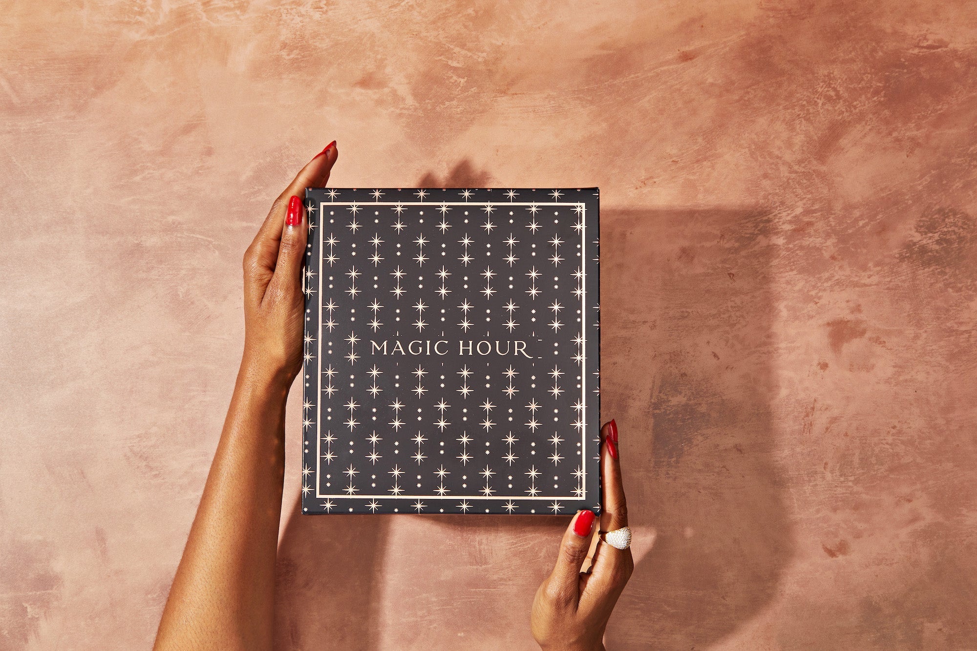 A pair of hands with red nails holds a black box with a starry pattern and the words "MAGIC HOUR" printed on it. The box is set against a textured, warm-toned background.