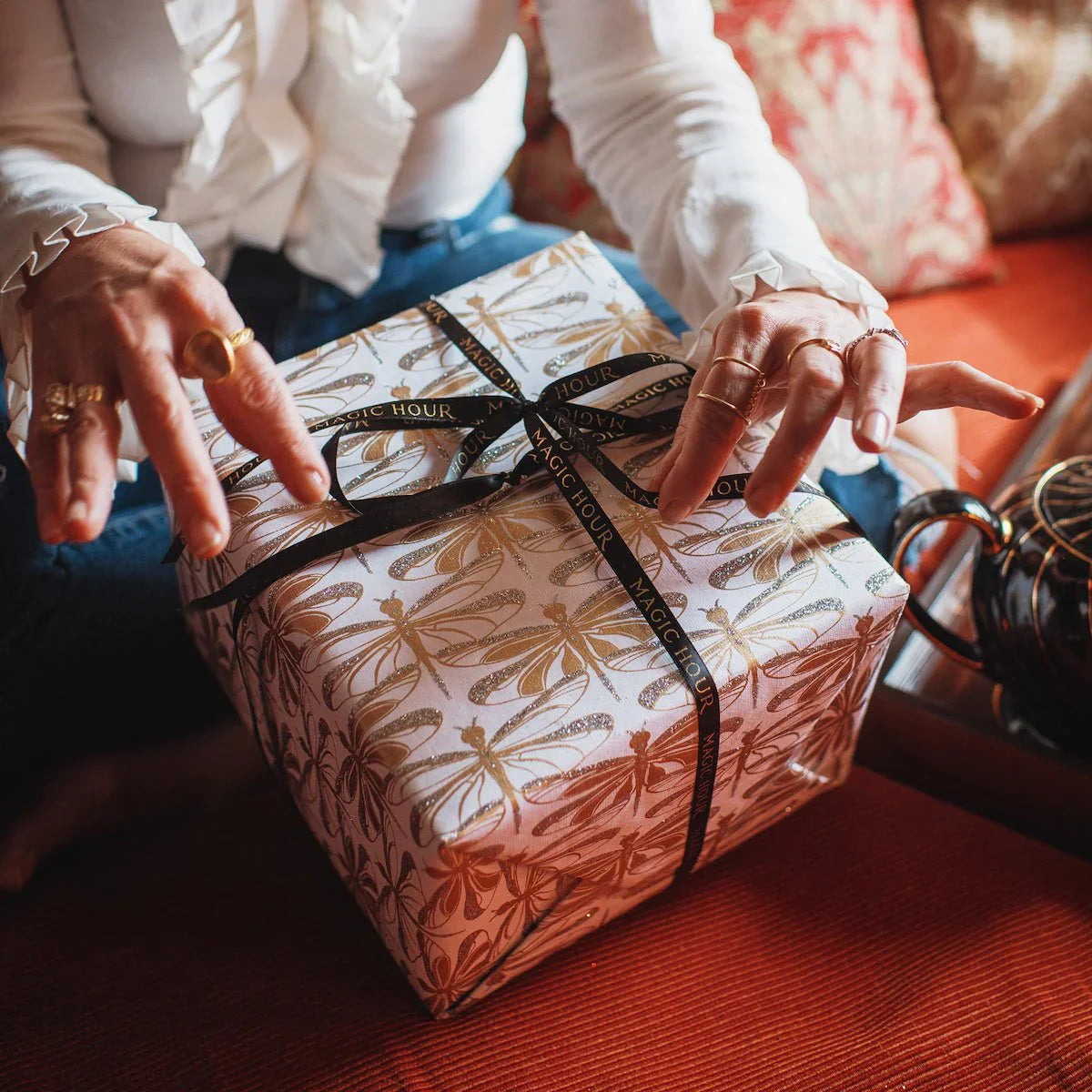 A person wearing rings and a white blouse sits on a couch, holding a wrapped gift box with decorative paper and a black ribbon. A teapot is nearby on the table.