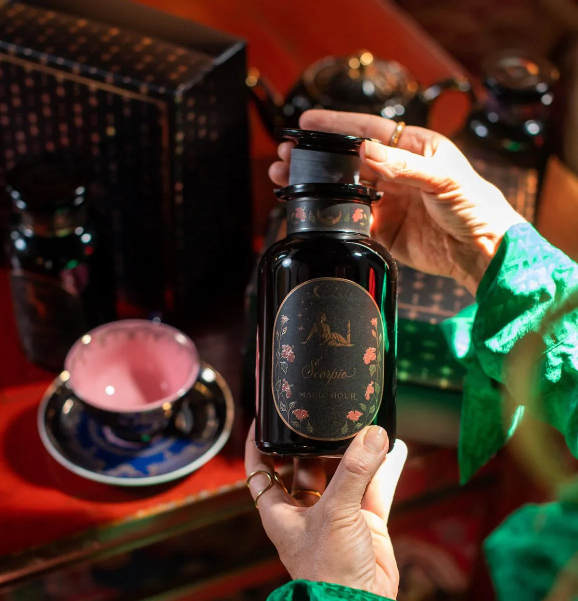 Hands holding a dark decorative bottle with a floral label, surrounded by a tea set and boxes on a wooden table. A pink teacup in a blue saucer is visible in the background.