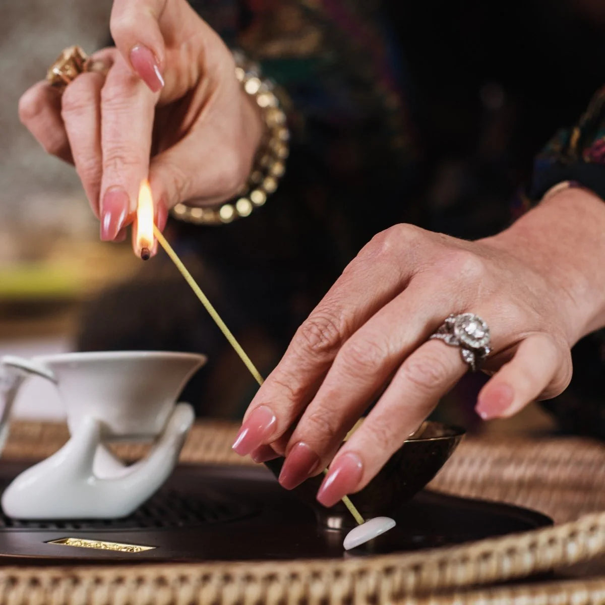 A close-up of hands with pink nails lighting an incense stick over a holder. The person wears multiple rings and a bracelet. A decorative teapot is in the background. The scene is set on a textured surface.