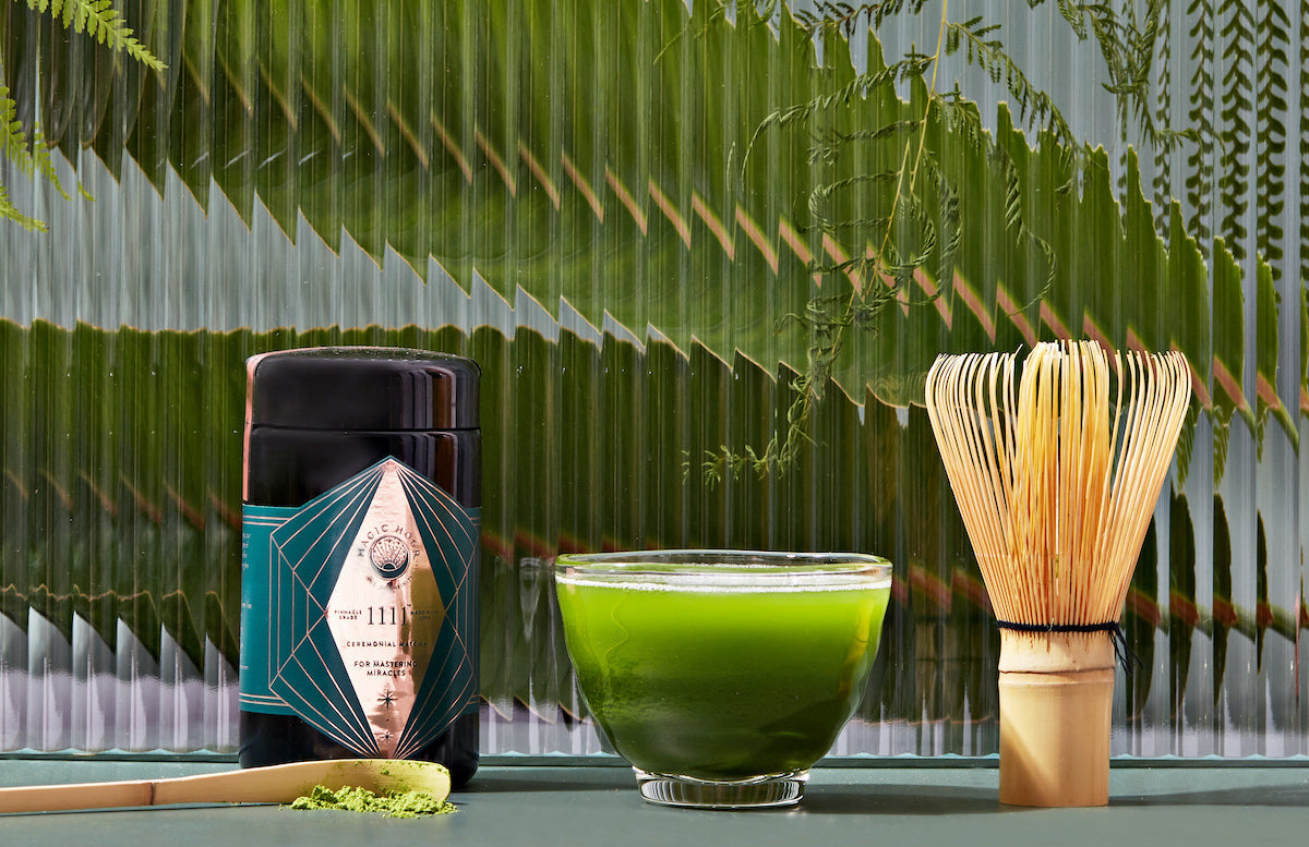 A jar of organic matcha sits next to a glass bowl filled with vibrant green matcha tea. A traditional bamboo whisk and a bamboo scoop with matcha powder are placed nearby. The background features lush green foliage and a textured glass surface.