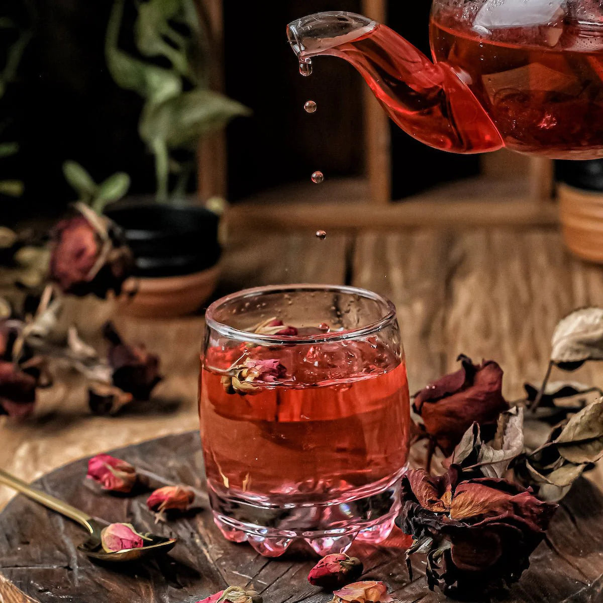 A glass teapot pours red tea into a clear glass with a wooden surface underneath. Dried rose petals are scattered around the board, with some inside the glass and on a spoon nearby. The background includes blurred plants in small pots.