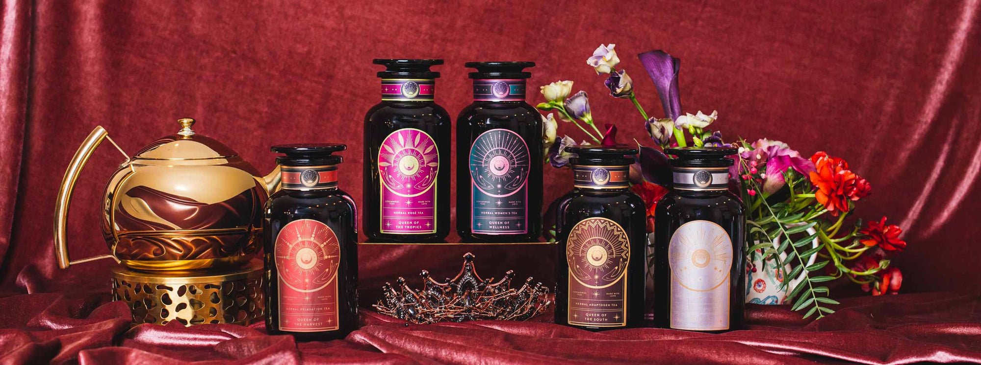 Five elegant tea canisters with intricate labels are displayed on rich red fabric, surrounded by a gold teapot, a jeweled crown, and vibrant flowers for a luxurious presentation.