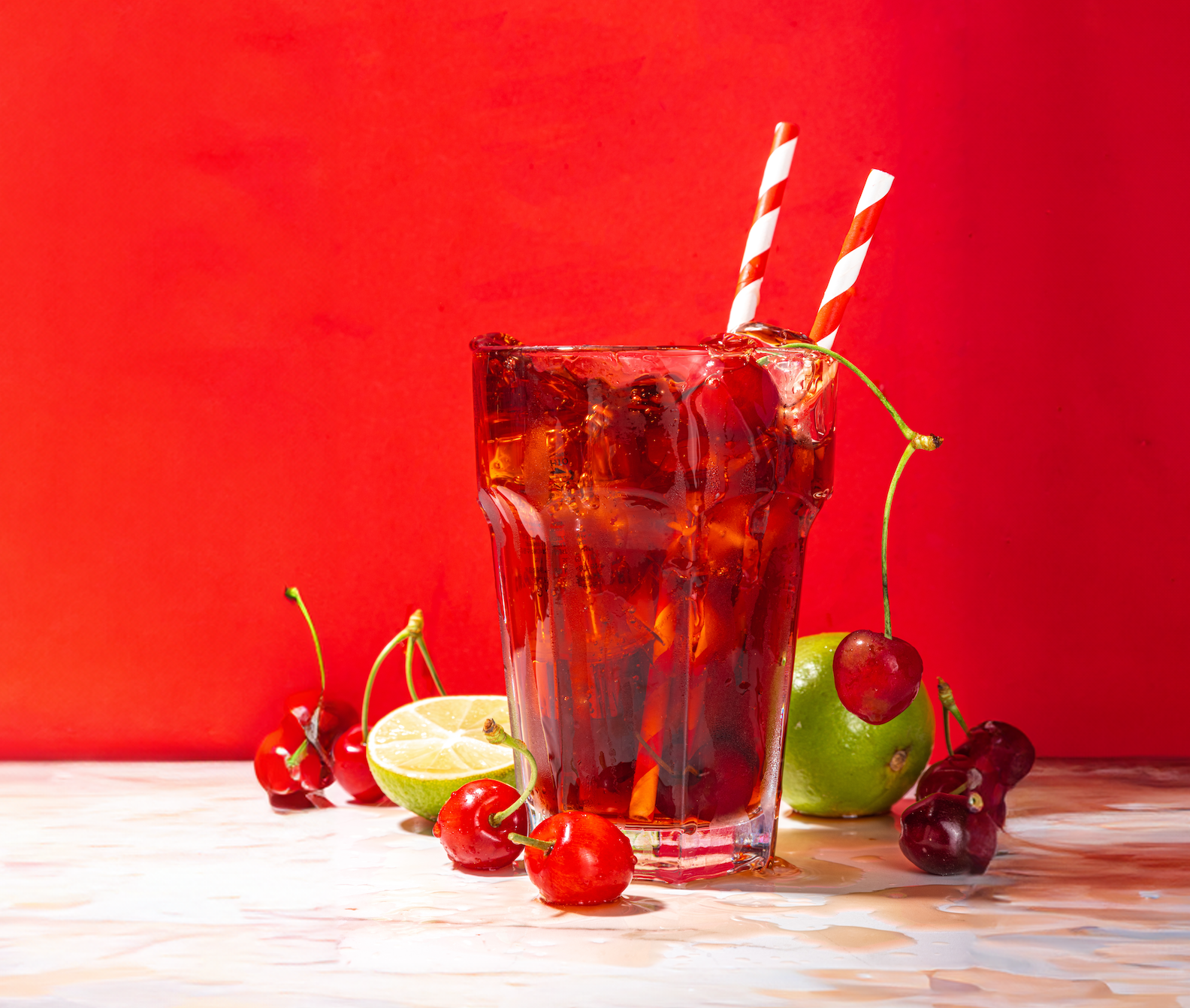 A glass of dark soda with ice and two red striped straws sits amidst fresh cherries, a lime half, and a green apple against a vibrant red background.