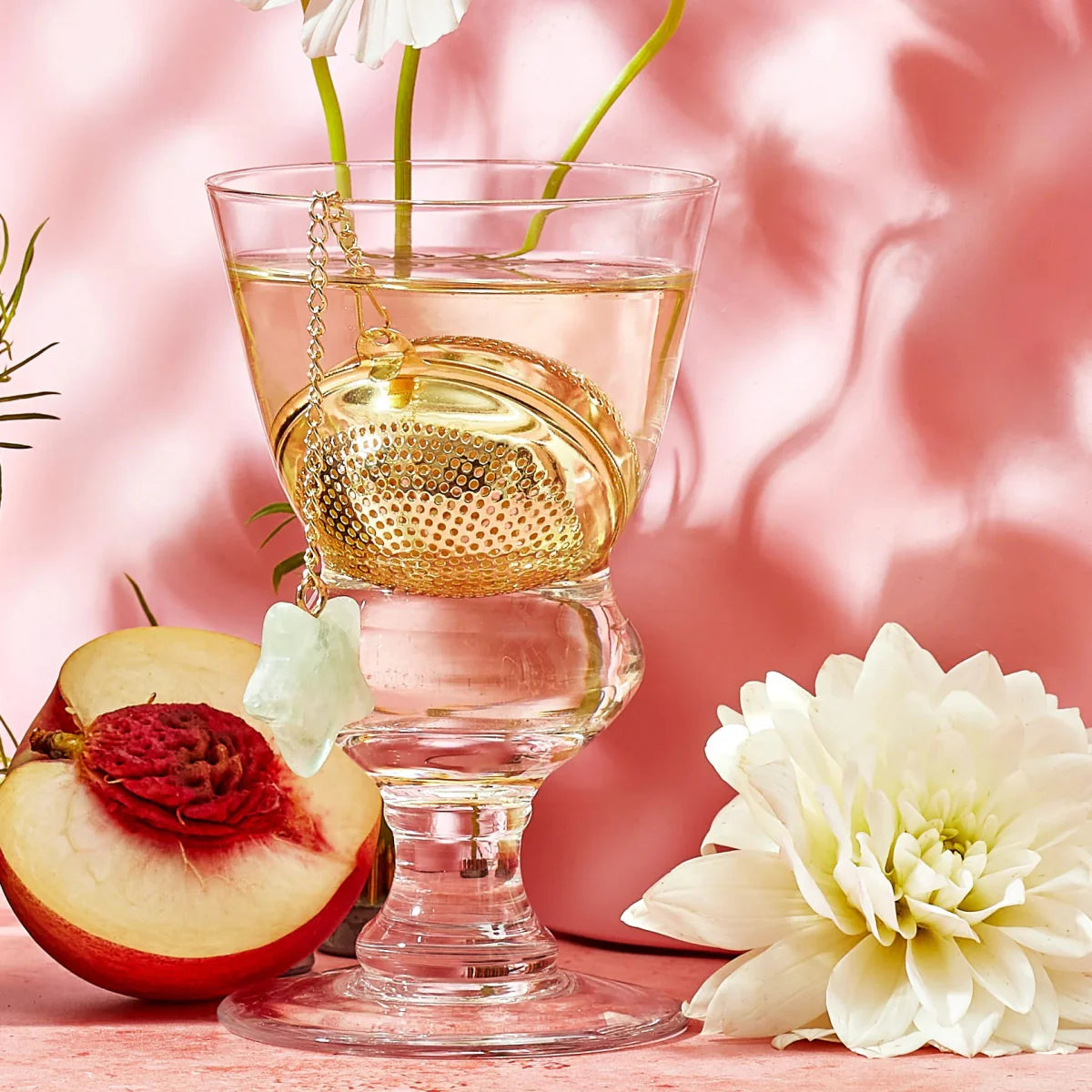 A glass of tea with a gold tea infuser is surrounded by a white chrysanthemum, a half-sliced peach, and pink shadows. Green stems of flowers are visible in the tea. The setting has a romantic tone.