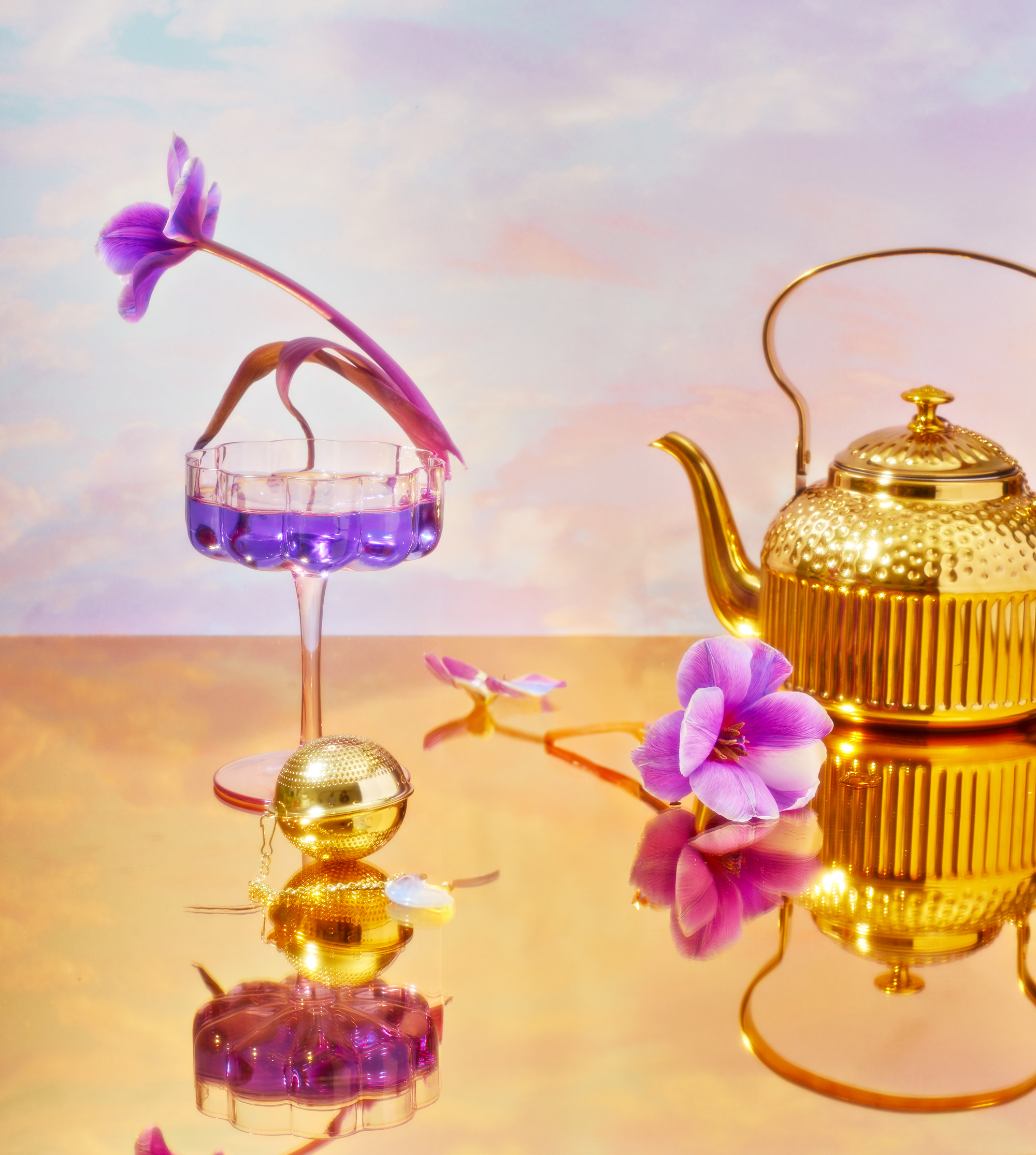 A gold teapot and a glass with purple liquid are on a reflective surface, surrounded by purple flowers, one nestled inside the glass. The scene evokes serene vibes and invites thoughts of exploring wellness with the School of Magic Tea Subscription Box - Traveler Jar.