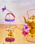 A gold teapot and a glass with purple liquid are on a reflective surface, surrounded by purple flowers, one nestled inside the glass. The scene evokes serene vibes and invites thoughts of exploring wellness with the School of Magic Tea Subscription Box - Traveler Jar.