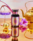 A vibrant still life composition showcases a golden teapot, a bottle labeled "1111: Seraphim's Dream Tea," a purple flower in a glass filled with purple liquid, and a tea strainer, all arranged against a reflective surface highlighted by gentle, colorful lighting.