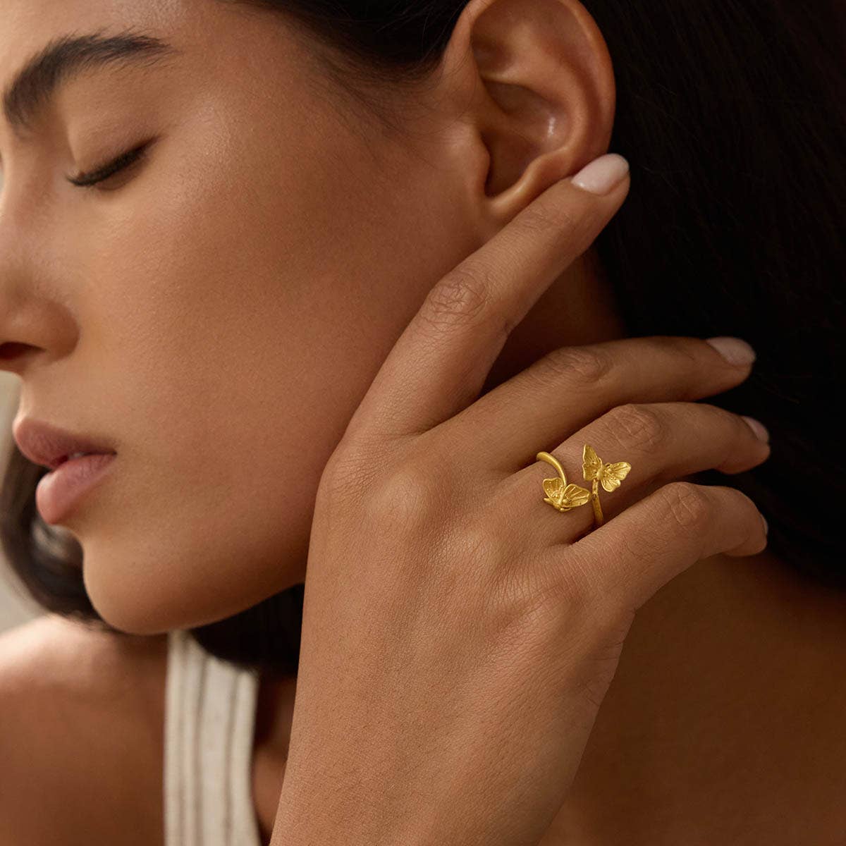 A woman with smooth skin and dark hair, eyes closed in calm, touches her face while wearing the Freedom to Soar Adjustable Butterfly Double Butterfly Ring. She pairs the elegant ring with a white top.