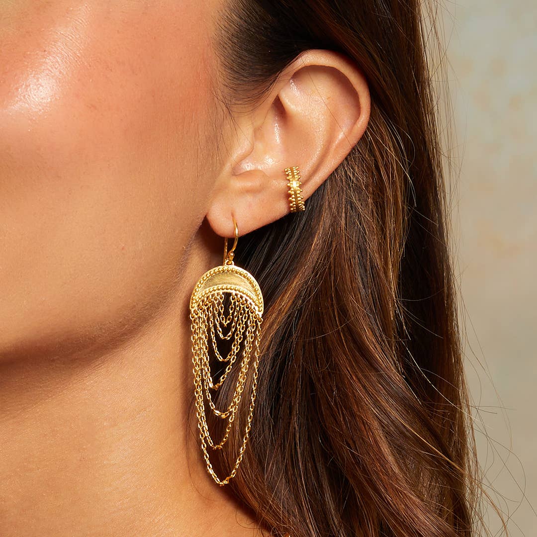 A close-up of a woman's ear showcases the Drape Chain Moon Earrings—18kt gold-plated earrings with layered chains hanging from a half-circle. Her brown hair is swept aside, revealing her smooth, glowing skin.