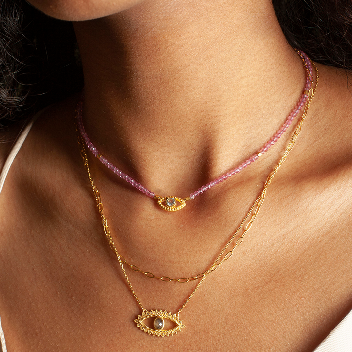 A woman in a white top wears three layered necklaces: a pink beaded strand, and two gold plated pieces—one being the Eye Blue Topaz Necklace with an eye-shaped pendant featuring blue topaz.