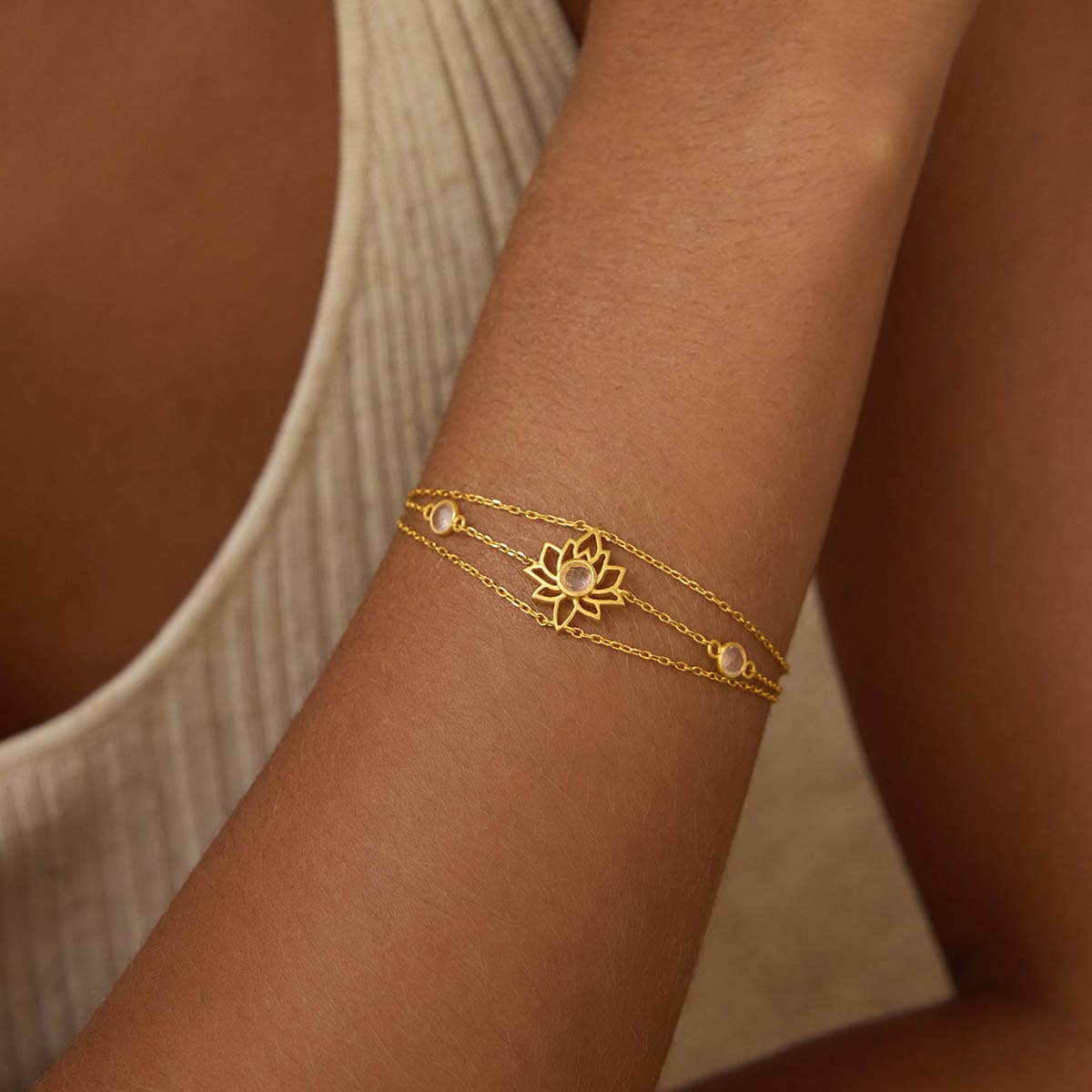 A person wears the Transformed by Love Lotus Bracelet—an 18k gold-plated double chain with a lotus flower charm and small white beads—on their forearm, paired with a light-colored sleeveless top.