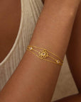 A person wears the Transformed by Love Lotus Bracelet—an 18k gold-plated double chain with a lotus flower charm and small white beads—on their forearm, paired with a light-colored sleeveless top.