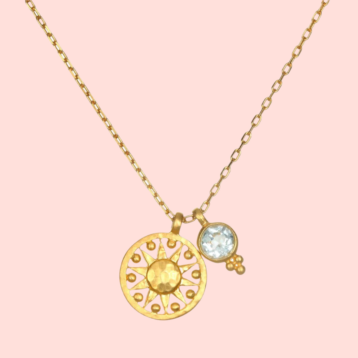 The Blue Topaz Sun Pendant Necklace features a gold-plated sun-shaped pendant with a textured center and dotted rays, paired with a small round blue topaz charm accented by three tiny gold beads for radiant style.