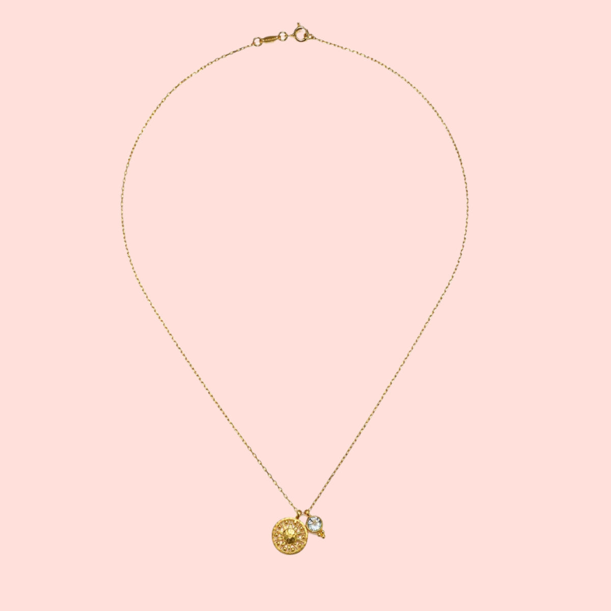 The Blue Topaz Sun Pendant Necklace features a delicate gold plated chain with a round, textured gold pendant and a small blue topaz gemstone charm, displayed on a white background.