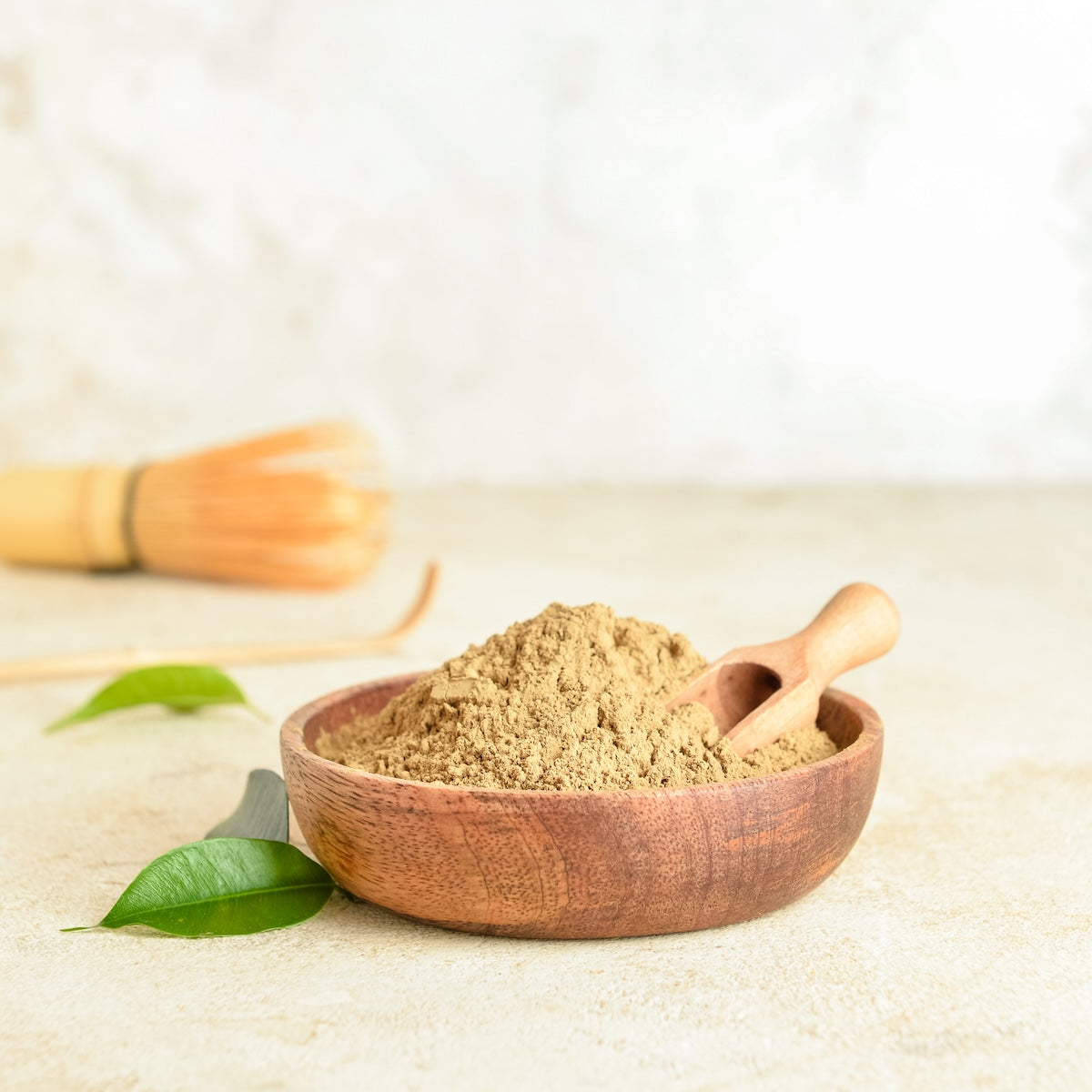 A wooden bowl of Hojicha powder and a small scoop sit on a light surface with green leaves, while a bamboo whisk blurs in the background, reflecting the comforting warmth of this low-caffeine Japanese green tea.