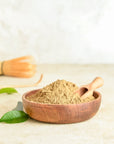 A wooden bowl of Hojicha powder and a small scoop sit on a light surface with green leaves, while a bamboo whisk blurs in the background, reflecting the comforting warmth of this low-caffeine Japanese green tea.