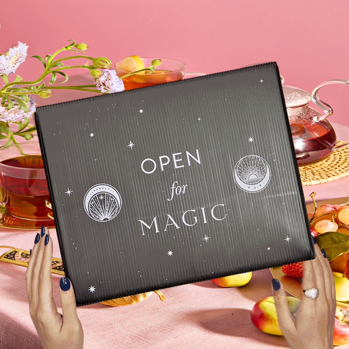 Hands holding a Super Spring Mystery Box over a table with spring teas, flowers, a glass teapot, tea cups, fruit assortment, and a whimsical pink background.