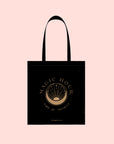 The Magic Hour Tote Bag is a black cotton tote bag featuring a metallic gold logo with a sunrise design. The words "MAGIC HOUR" and "TIME for INFINITY" gracefully encircle the emblem, set against a light peach background.