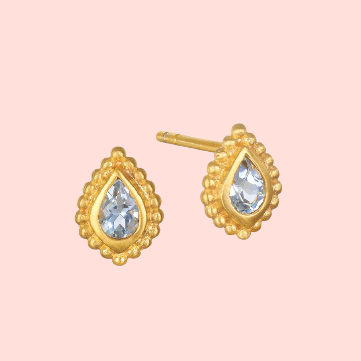 The Inner Light Blue Topaz Pear Stud Earring features a pear-shaped design with a clear blue topaz center, surrounded by a textured gold border and small round beads, inspired by delicate lotus motifs.