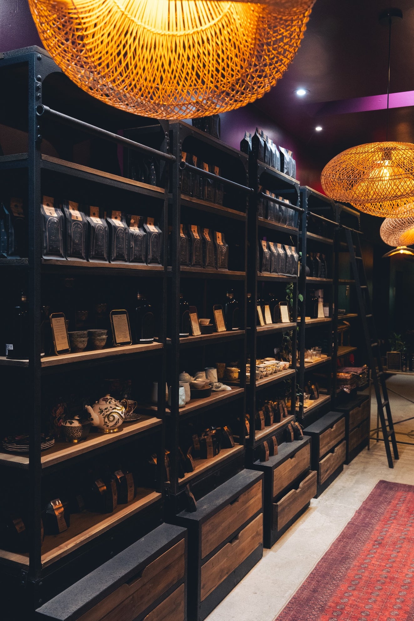 A cozy store with dim, warm lighting featuring tall black shelves filled with an assortment of jars, teapots, and various items. Wooden drawers at the bottom of the shelves and woven chandelier lights hanging from the ceiling add to the ambiance.