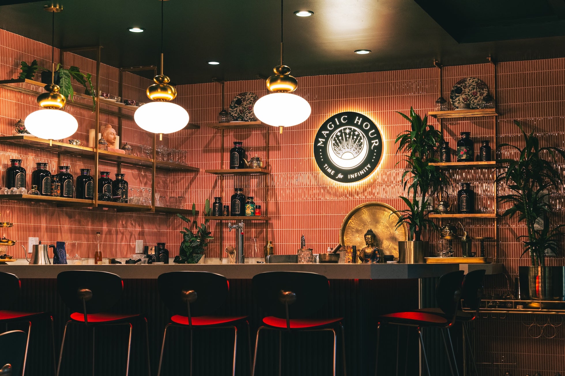 A stylish bar with a neon sign reading "Magic Hour" on the back wall. The decor features pink tiles, gold accents, and lush green plants. The bar counter is adorned with glassware and bottles, while modern hanging lights illuminate the red-cushioned bar stools.
