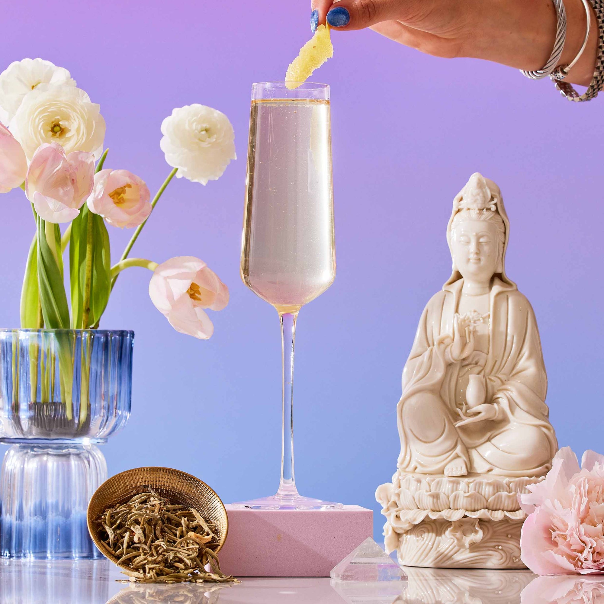 A hand garnishes the French 75: Lemon Sparkling Wine White Mocktail Tea for Cool, Calm Radiance in a flute, with pink flowers in a blue vase, Silver Needle white tea in a gold dish, a white statue, and a pink flower on a purple gradient backdrop.