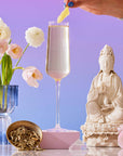 A hand garnishes the French 75: Lemon Sparkling Wine White Mocktail Tea for Cool, Calm Radiance in a flute, with pink flowers in a blue vase, Silver Needle white tea in a gold dish, a white statue, and a pink flower on a purple gradient backdrop.
