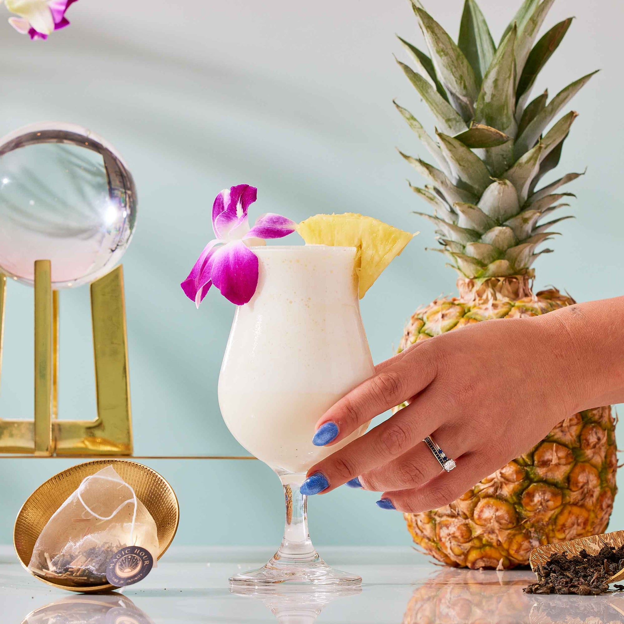 A hand with blue nails holds a pina colada garnished with a pineapple slice and a purple flower—capturing the essence of summer refreshment. Nearby, a whole pineapple, a crystal ball, and a shimmering gold luxury tea infuser rest atop the glossy surface, evoking the elevated delights found in The Best of Summer Poolside Sips Box.