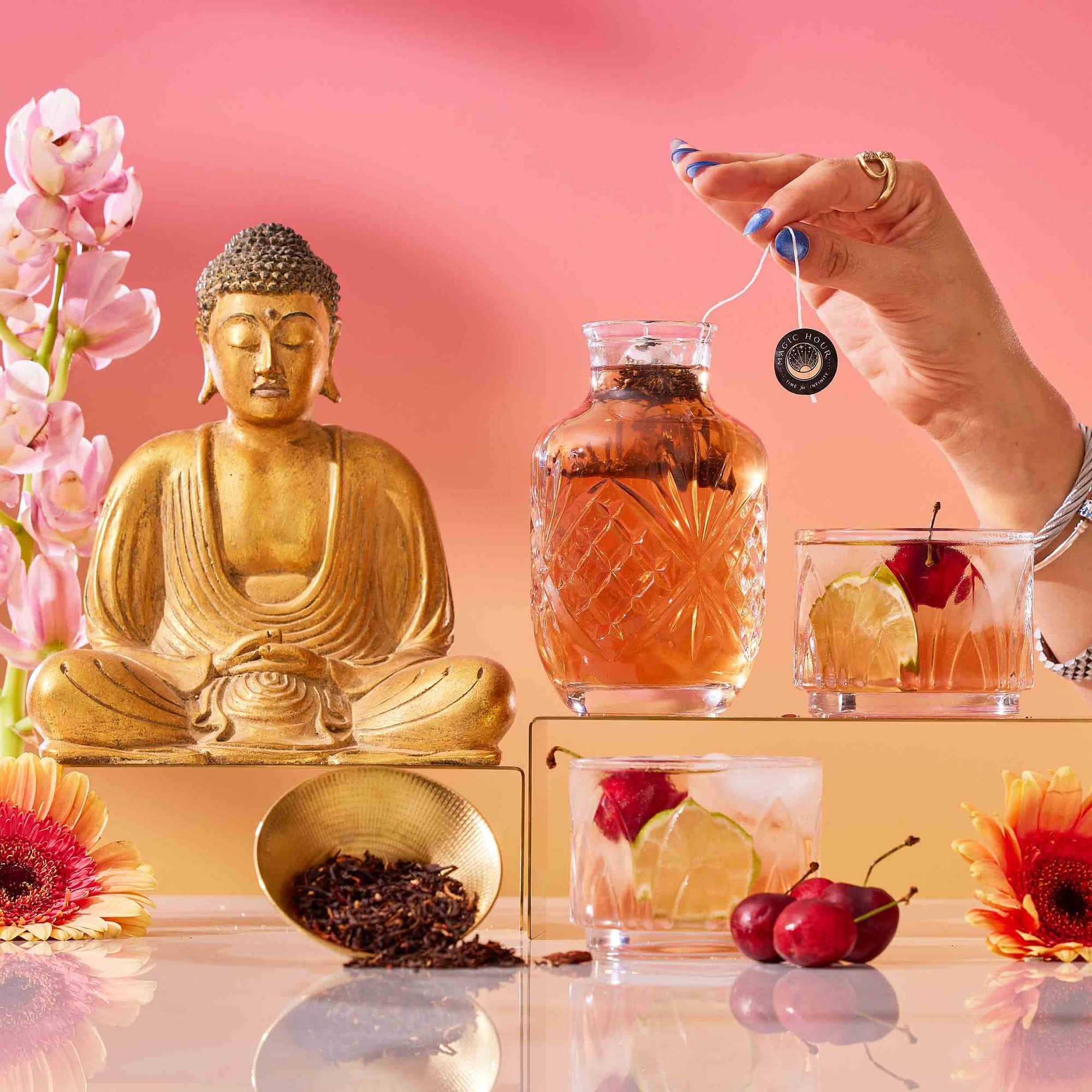 A hand holds a tea bag above a glass teapot beside a Buddha statue, flowers, bowls of Mai Tai: Black & Puerh Tropical Mocktail Tea, fruit cocktails, and cherries—all on a reflective surface with a pink-orange backdrop.