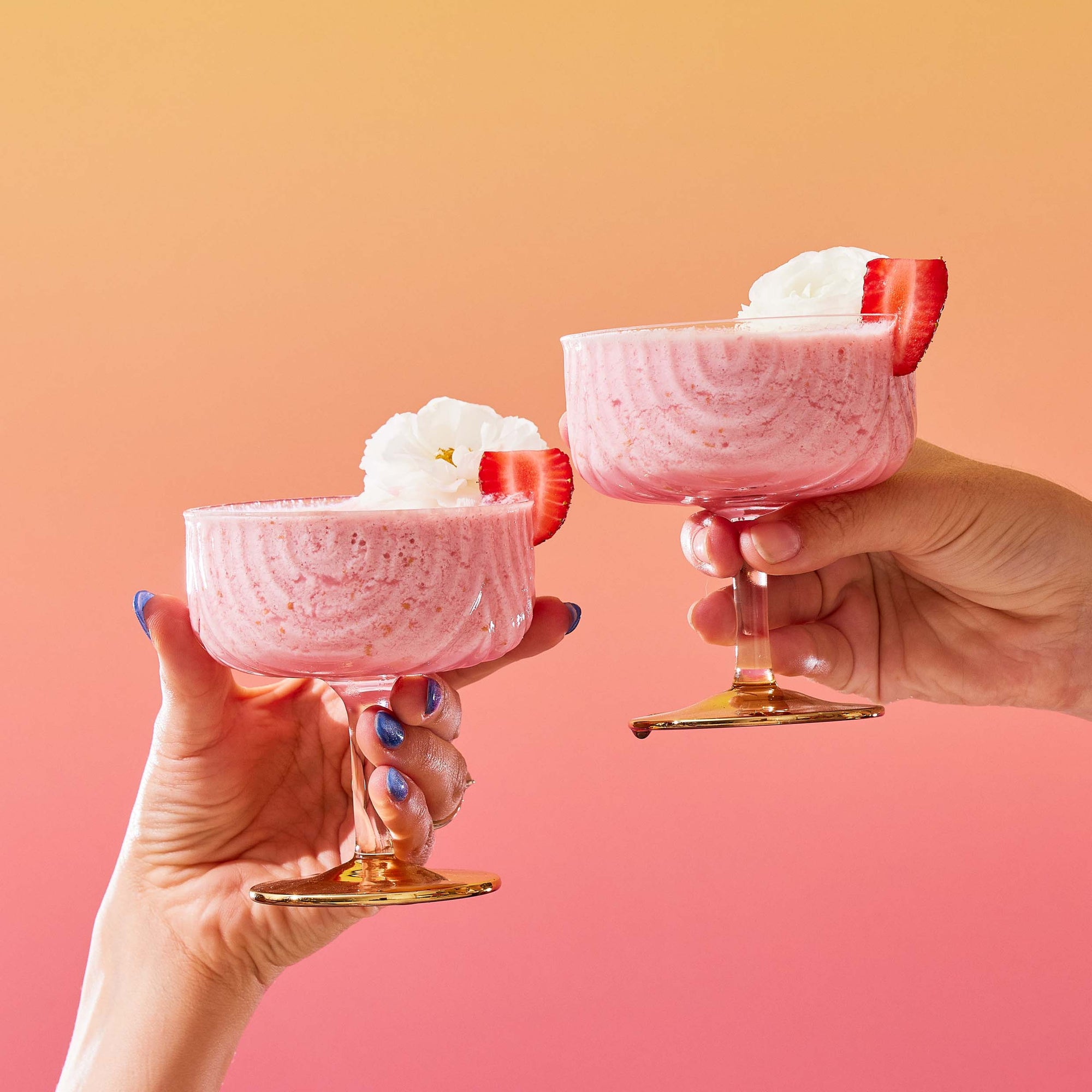 Two hands holding pink strawberry milkshakes in coupe glasses, topped with whipped cream and a strawberry slice, set against a gradient orange-to-pink background—perfect for enjoying summer sips or pairing with treats from The Best of Summer Poolside Sips Box.