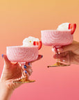 Two hands holding pink strawberry milkshakes in coupe glasses, topped with whipped cream and a strawberry slice, set against a gradient orange-to-pink background—perfect for enjoying summer sips or pairing with treats from The Best of Summer Poolside Sips Box.
