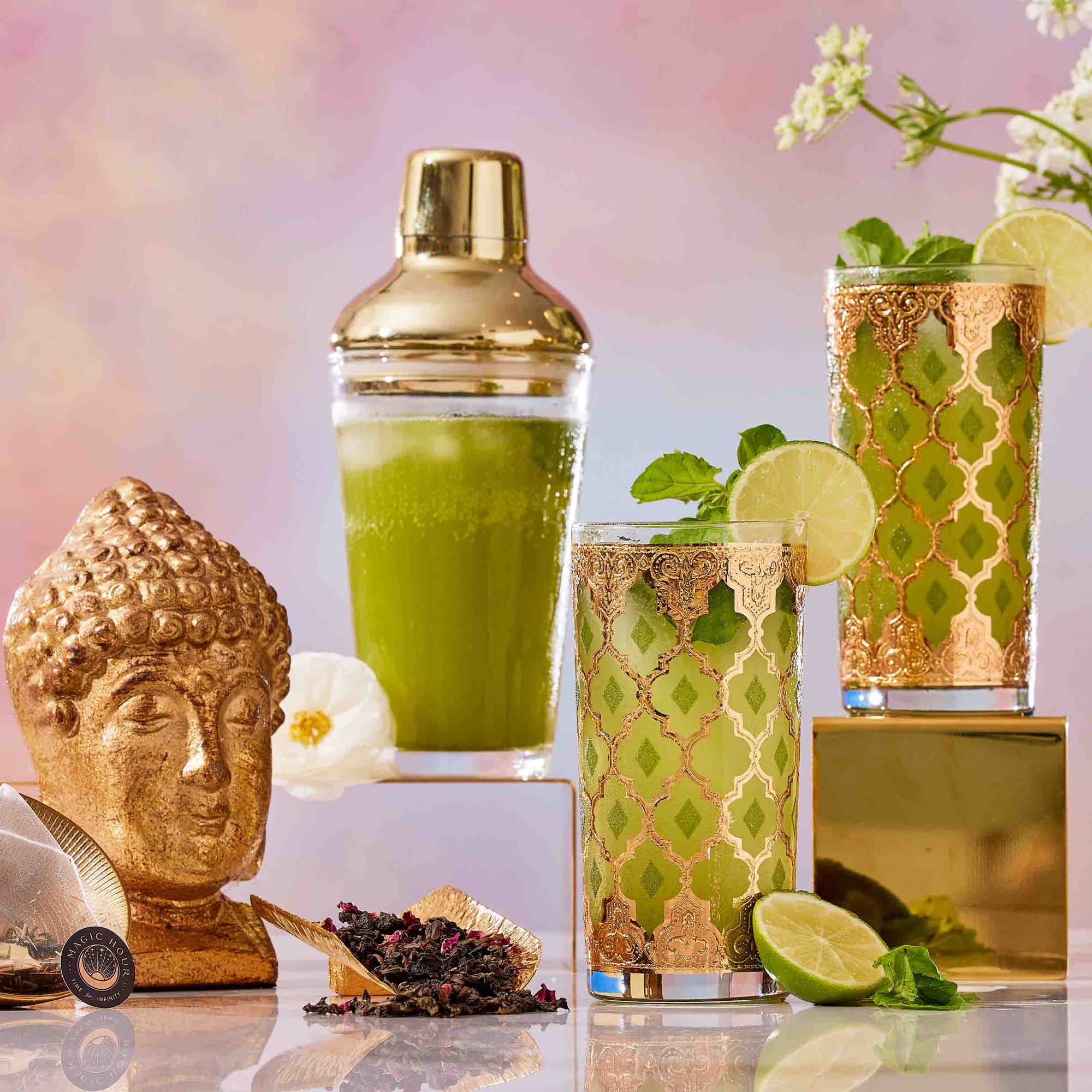 Two ornate gold glasses filled with refreshing green drinks, garnished with lime and mint, rest beside a gold Buddha head, cocktail shaker, white flower, loose tea, and a gold grater—all set against a pastel backdrop. This elegant arrangement captures the essence of The Best of Summer Poolside Sips Box, inspiring luxurious tea moments and perfect summer gifting ideas.