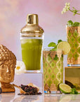 Two ornate gold glasses filled with refreshing green drinks, garnished with lime and mint, rest beside a gold Buddha head, cocktail shaker, white flower, loose tea, and a gold grater—all set against a pastel backdrop. This elegant arrangement captures the essence of The Best of Summer Poolside Sips Box, inspiring luxurious tea moments and perfect summer gifting ideas.