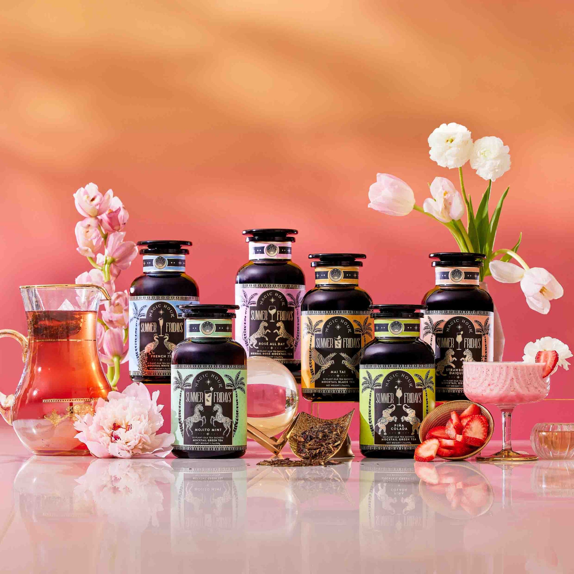 A set of "Summer Fridays: Wellness Mocktail Tea Bundle" black tea jars is arranged with glass teapots, flowers, sliced fruit, and mocktail teas on a reflective surface against a soft pink and orange gradient background.