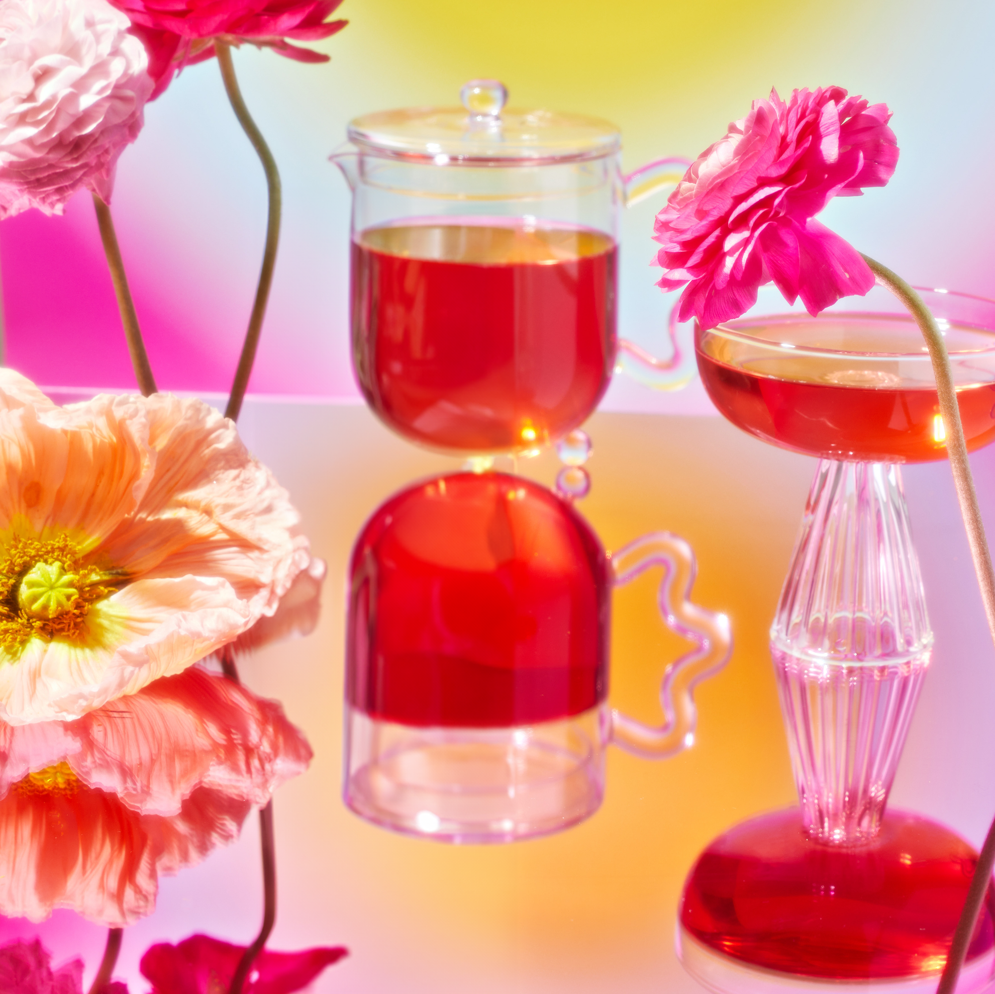 A vibrant image showcases a glass teapot and cocktail glass with red liquid on a reflective surface. Bright pink and orange flowers, part of the School of Magic Tea Subscription Box - Traveler Jar, surround the scene against a colorful gradient background.