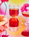 A vibrant image showcases a glass teapot and cocktail glass with red liquid on a reflective surface. Bright pink and orange flowers, part of the School of Magic Tea Subscription Box - Traveler Jar, surround the scene against a colorful gradient background.