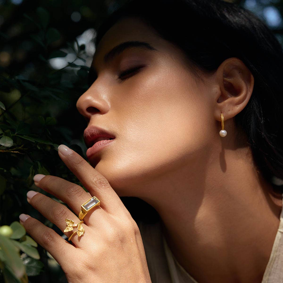A woman with closed eyes poses near greenery, wearing gold jewelry highlighted by soft sunlight, including pearl hoop earrings and the Freedom to Soar Adjustable Butterfly Double Butterfly Ring.