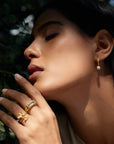 A woman with closed eyes poses near greenery, wearing gold jewelry highlighted by soft sunlight, including pearl hoop earrings and the Freedom to Soar Adjustable Butterfly Double Butterfly Ring.