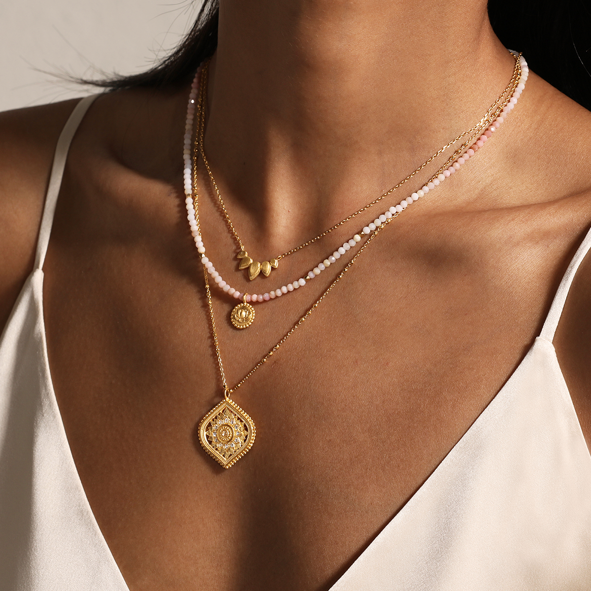 A woman in a white spaghetti strap top wears three layered necklaces: a pink and white beaded choker, a gold leaf charm, and an 18” White Topaz Lotus Medallion Necklace.