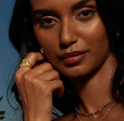 A woman with medium skin tone and dark hair poses with her hand near her face, wearing the Emerald Radial Evil Eye Ring set of 3, along with gold earrings and a necklace, against a dark blue background.