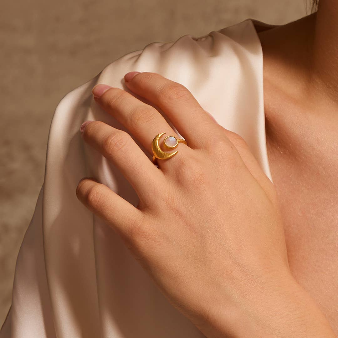 A hand rests on a shoulder, showcasing the Cradled in Light Moonstone Ring with a gold-plated crescent design and round white stone, set against a soft beige fabric background.