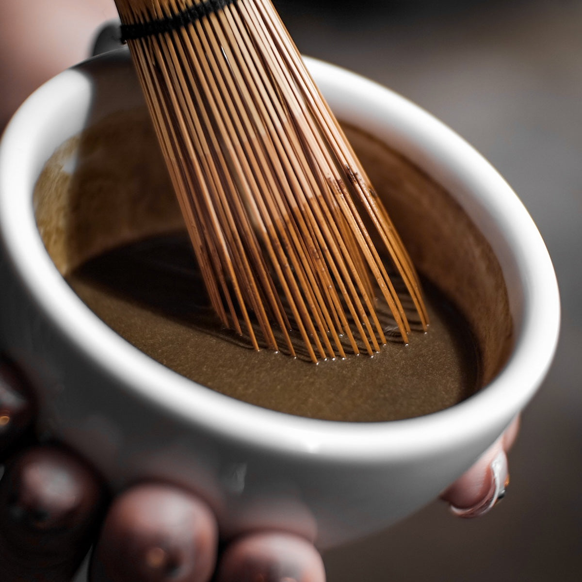 A hand holds a white bowl of frothy Hojicha, being whisked with a traditional bamboo whisk.