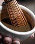 A hand holds a white bowl of frothy Hojicha, being whisked with a traditional bamboo whisk.