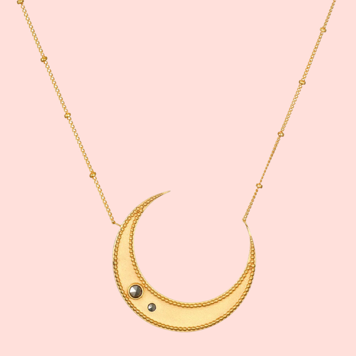 The Satya Jewelry Eternal Guidance Moon Necklace features a gold-plated crescent moon with two small dark pyrite stones on the lower left, hanging from a delicate beaded gold chain.