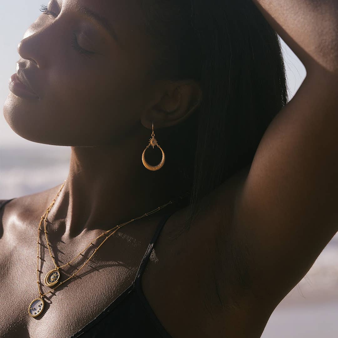 A woman with dark skin poses outdoors, eyes closed and sunlight highlighting her face, wearing a black top and the Stellar Sisterhood Moonstone Hoops with layered goddess jewelry.