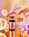 The vibrant display features the School of Magic Tea Subscription Box in a colorful bottle set among elegant orchids and calla lilies. A pink-nailed hand gently touches the glass jar with golden liquid, reminiscent of wellness tea, against a warm, gradient background.