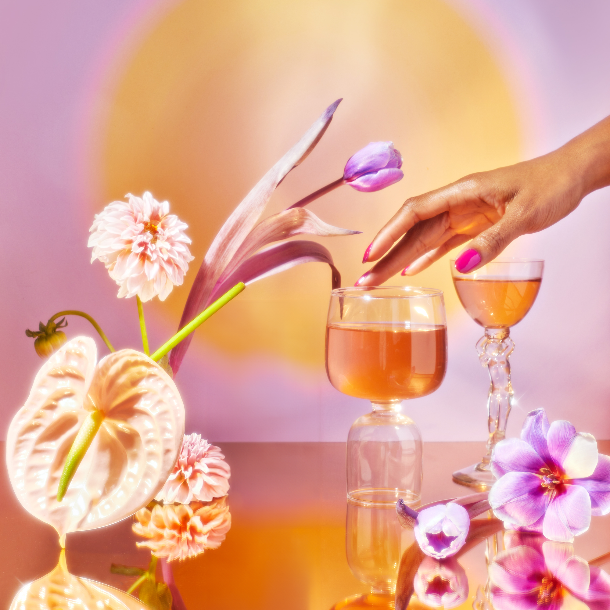 Pink nails gently caress a glass of pink liquid, possibly from the School of Magic Tea Subscription Box - Traveler Jar, resting on a reflective surface. Vibrant flowers and another filled glass surround the scene, set against a warm backdrop—a perfect mindfulness moment.