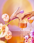 Pink nails gently caress a glass of pink liquid, possibly from the School of Magic Tea Subscription Box - Traveler Jar, resting on a reflective surface. Vibrant flowers and another filled glass surround the scene, set against a warm backdrop—a perfect mindfulness moment.