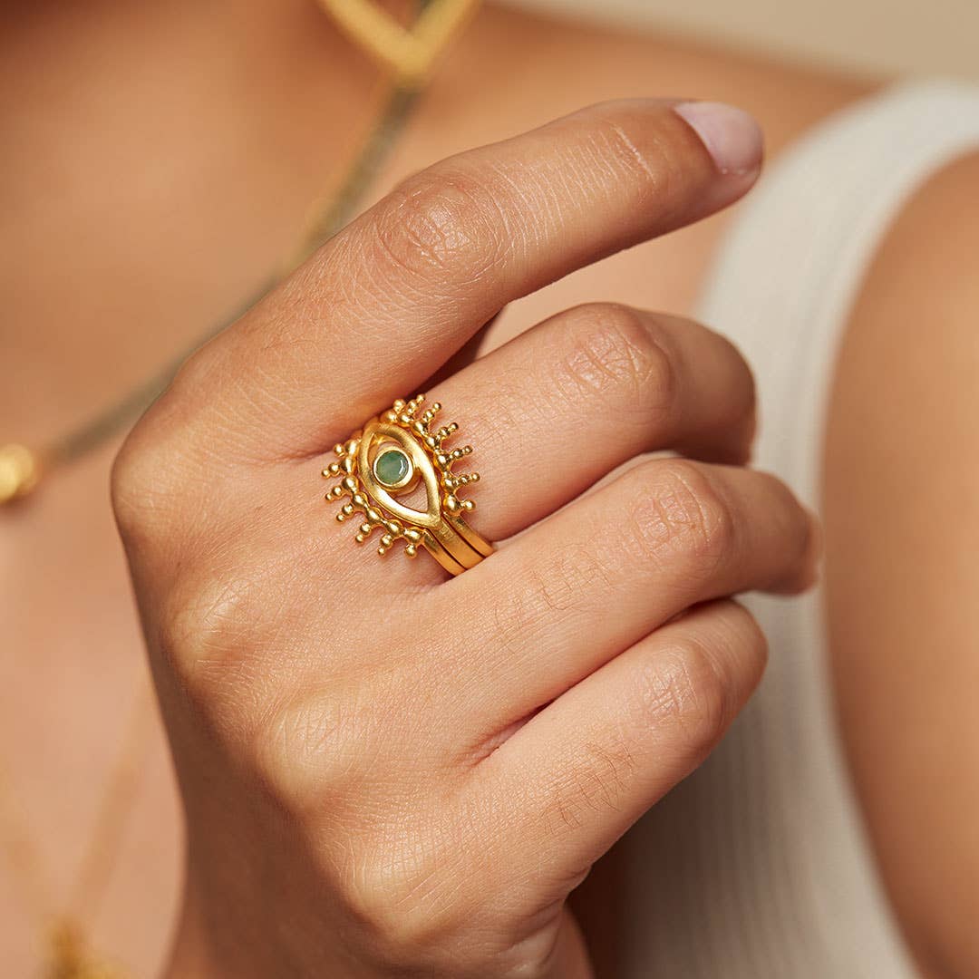 A hand showcases the Emerald Radial Evil Eye Ring set of 3, gold plated with green gemstones, worn on the ring finger. A white sleeveless top and a gold necklace are also visible in the background.